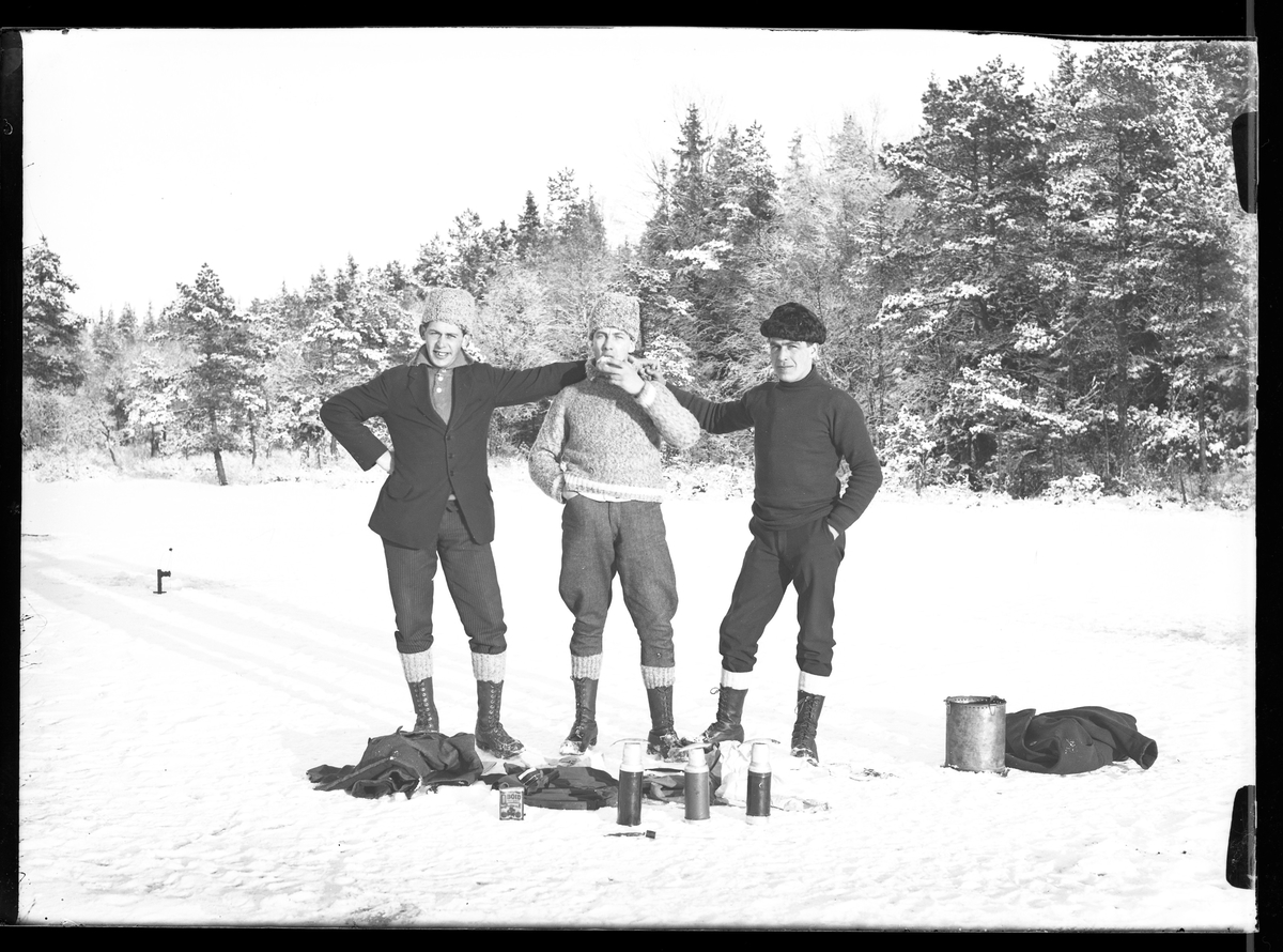 Tre vinterklädda män håller om varandra på isen. Framför dem ligger termosar och utrustning. Mannen i ljus pälsmössa och kavaj är troligen Hugo Larsson. I fotografens egna anteckningar står det "Angelfiske på Mörke". Om det är Lilla eller Stora Mörken framgår inte.