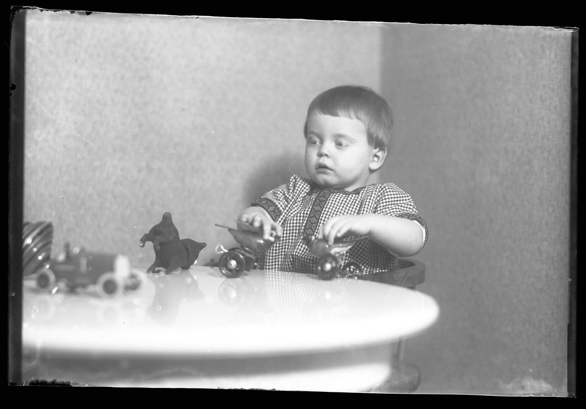 En liten pojke i rutig klänning sitter vid ett bord. På bordet syns en tygkatt, en bil, en snurra och pojken håller i två plåtfåglar. I fotografens egna anteckningar står det "Gust. Claessons pojke"