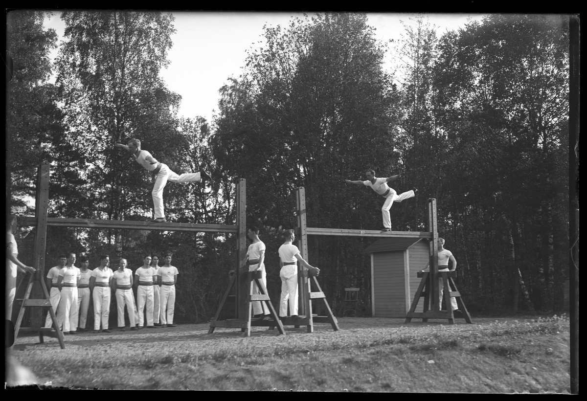 Alingås Gymnastikförenings manliga trupp har uppvisning. Gymnasterna har vita dräkter och två av dem balanserar på varsin bom. I fotografens egna anteckningar står det "A.G.F.s uppvisningar i Parken".
