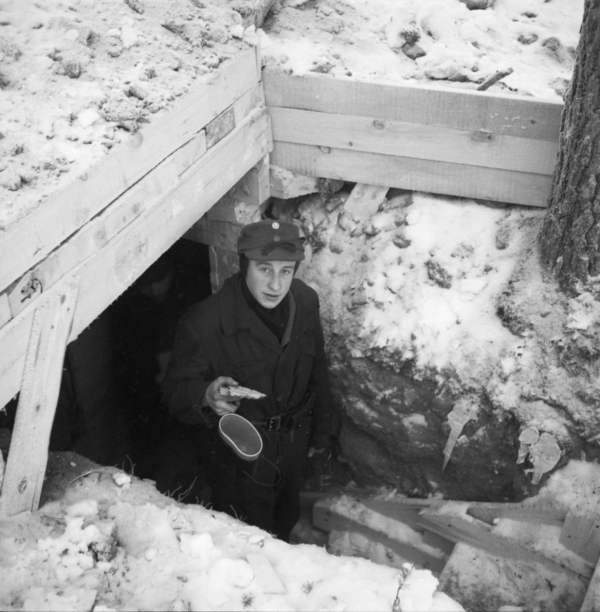 En militär står i trappen till ett markvärn efter utspisning. I handen håller han bröd och matkärl 'snuskburk'. Vid Svenska frivilligkåren i Finland, F 19.