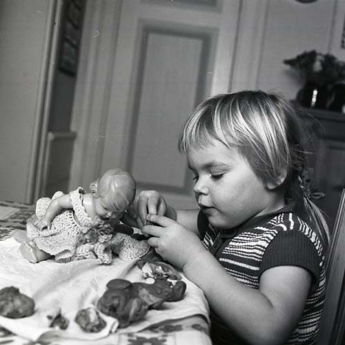 En flicka sitter vid ett bord och klär på en docka, 1972.