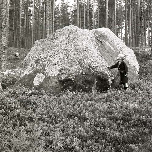 En pojke står intill en sten som är täckt av lav upptill. Pojken håller en hand mot stenen, 1985.
