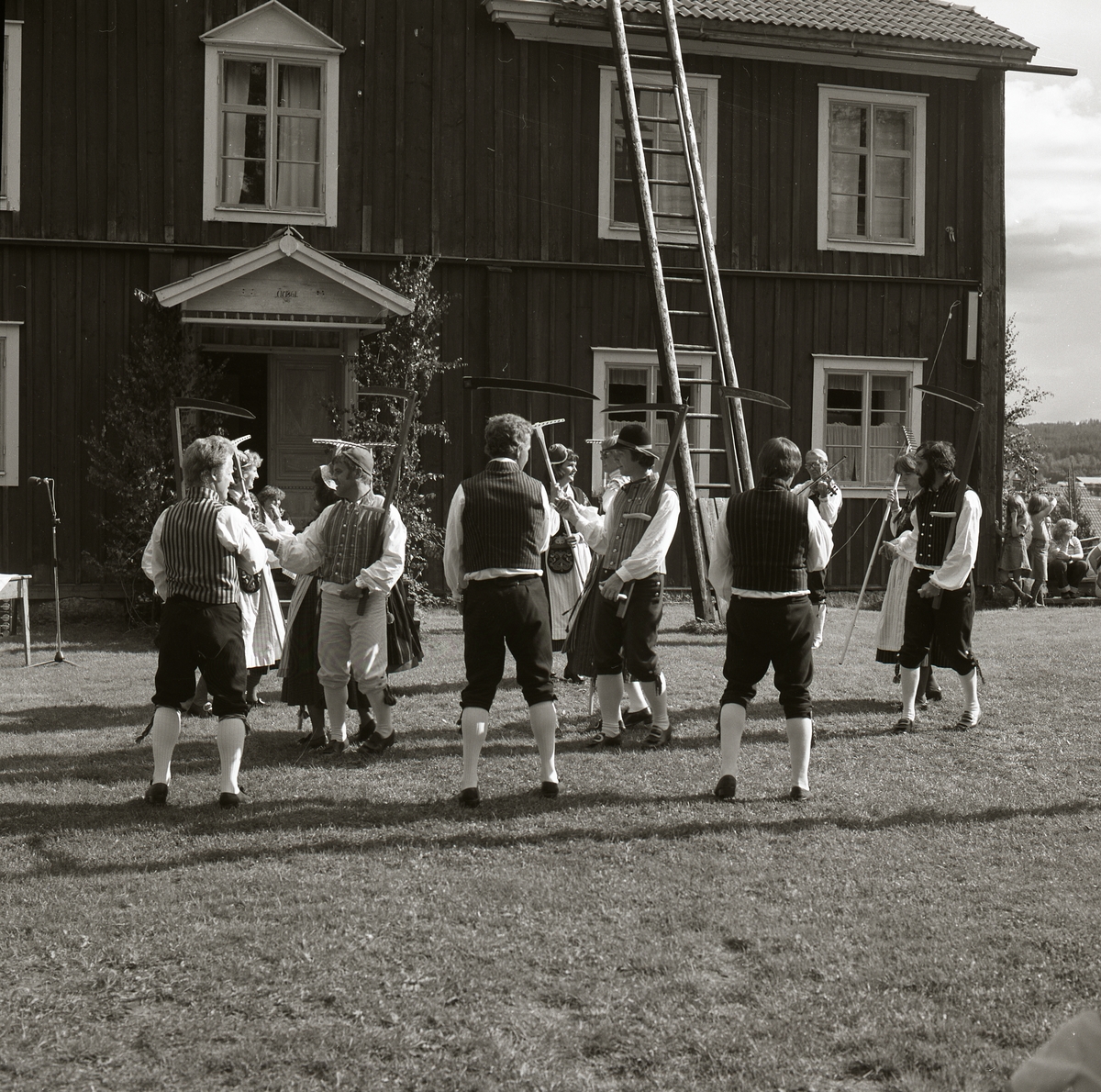 Några människor iförda hembygdsdräkter utför en dansuppvisning på gräsmattan utanför ett timrat hus under Rengsjöfesten, 16 juli 1978.
