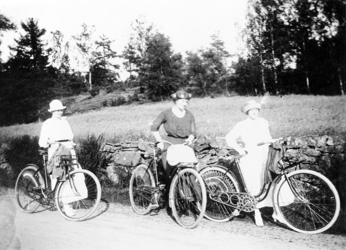 3 systrar (Jansson) står med var sin cykel, iklädda klänning och hatt, på en landsväg i deras levnadsort, Hjälmared. Deras far arbetade som sågverksarbetare.