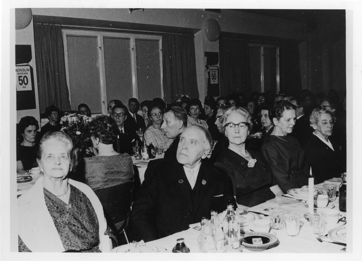 Överblicksbild inifrån en festlokal med män och kvinnor vid tre långbord. Konsum i Alingsås firar 50 år, (1915-1965), festen hålls i en okänd lokal.