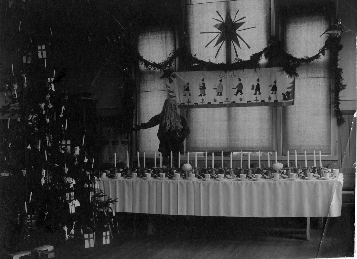 Bilden visar ett uppdukat långbord med kaffeporslin och ljusstakar. Bakom bordet en dekorativ jultomte, väggbonad med texten "GOD JUL" och en stor julstjärna. Till vänster ses en utsmyckad julgran med flaggor och ljus.
"Syföreningens" fest för barn "från mindre bemedlade föräldrahem". I början av 1900-talet.