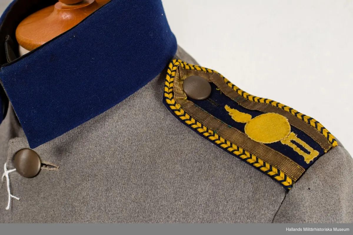 Vapenrocken är av grått kläde, är enradig med sex stycken bronserade, räfflade knappar, har ståndfällkrage (överkrage av blått kläde) är försedd med axelklaffar och chevron samt bär igenkänningstecken och skoltecken på vänster ärm (samtliga av blått kläde).  föreställande en granat samt romerska siffran II (2). Axelklaffarna har blå matta med emblem i gult kläde, guldgalon och gulblått beläggningssnöre längs med kanterna Ridbyxan är av grått kläde med blå klädlist längs med sömmarna på utsidan av benet och är i modellen avsmalnande från knät och nedåt.