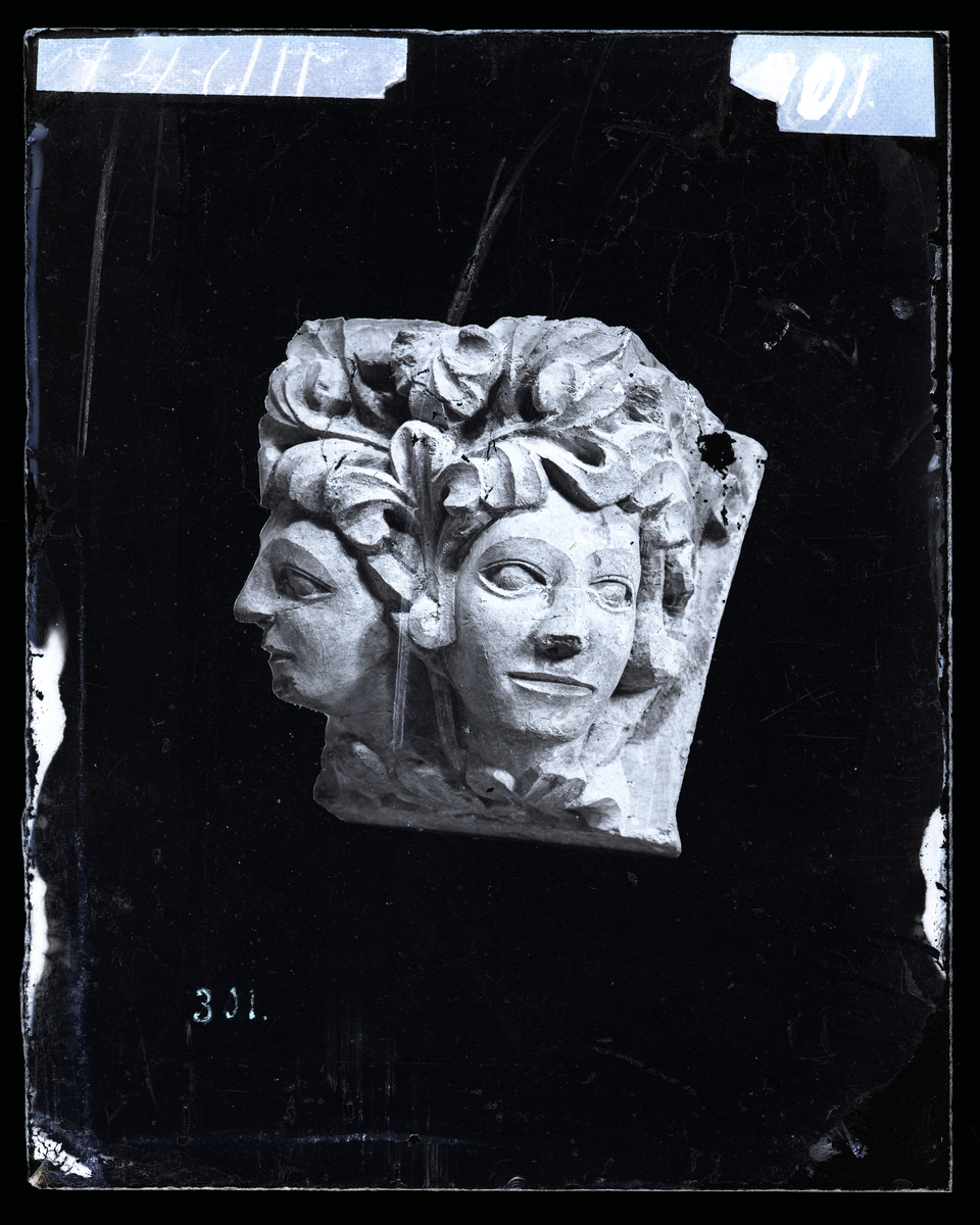Skulptur fra Nidarosdomen. En konsoll med to hoder. Ansiktene har utskårede øyne uten pupiller og bladverk i stedet for hår. Trolig en del av en buegesims rundt koret utvendig.