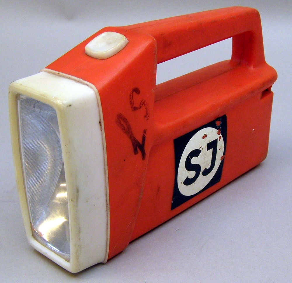 Ficklampa i orange och vit plast. Rektangulär form, med handtag längs större delen av ovansidan. En på- och avstängningsknapp av vit plast bredvid handtaget. Drivs med batteri. På varje långsida ett klistermärke med SJ:s logotyp som användes på 1960-70-och 80-talen. Med tuschpenna är skrivet för hand: "SJ" med skrivstil. 
Ficklampan är enligt uppgift en tämligen ovanlig sort inom järnvägsvärlden som troligtvis användes under 1970-talet.