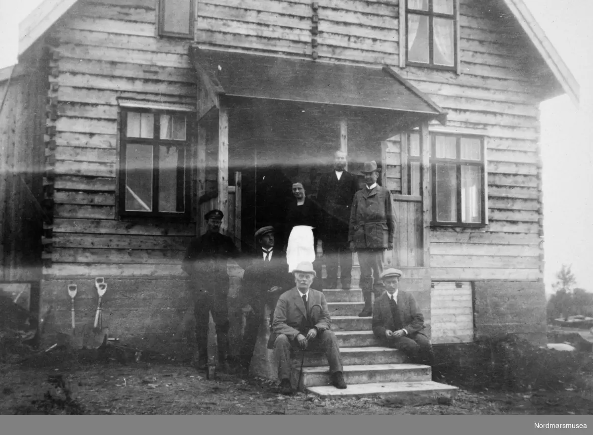 Foto av en gruppe menn og en kvinne samlet på trappen til et bolighus. Et bilde fra en samling fotografier med bureisingsfelt i Norge. Originalmaterialet tilhører Norsk myrmuseum i Smøla kommune. Fra Nordmøre museums fotosamlinger.