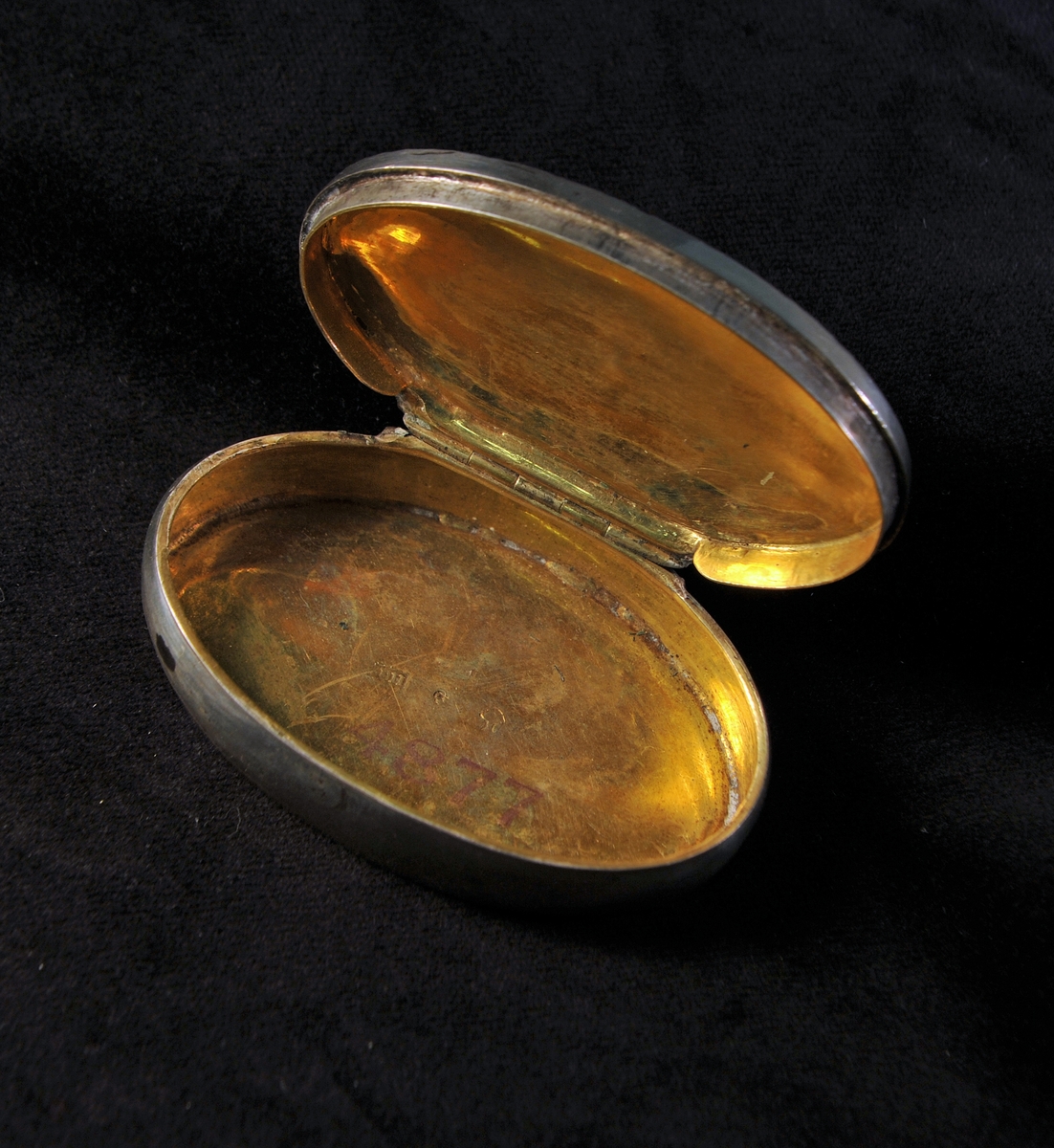 Oval, något buktande dosa av silver. Locket dekorerat med filigranarbete och 7 infattade stenar av rött glas. Invändigt förgylld. Stämplad invändigt i botten.