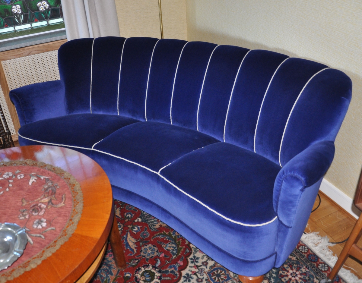 Sofa trukket med blå tekstil og hvite snorer. Sofaen har en buet form.