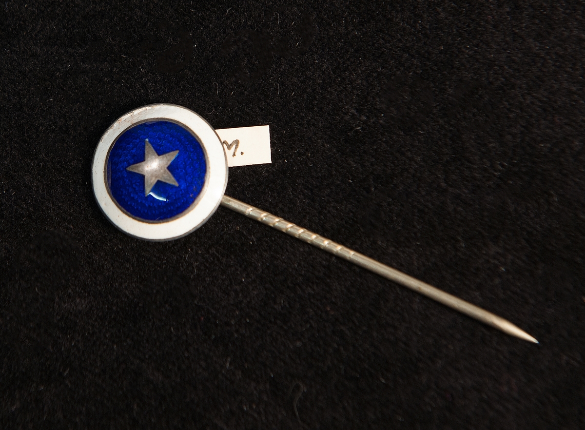 Kråsnåll i form av rund platta med emalj i blått och vitt, samt stjärna av silver(?) (tenn?) i mitten.