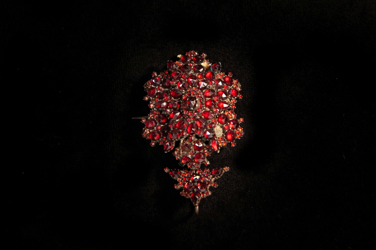 En brosch av metall, i form av en sexuddig stjärna besatt med slipade röda granater. Försedd med ett trekantigt hänge, besatt med slipade granater