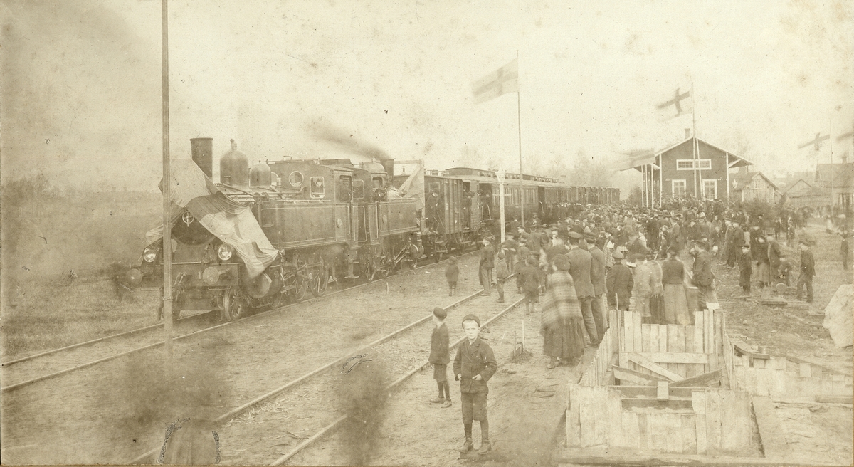 Invigningståget vid Tidaholms station 1906. Ångloken Vulcan och Tidaholm som är draperade med flaggor drar festtåg till Tidaholms station där en stor folkmassa väntar.
