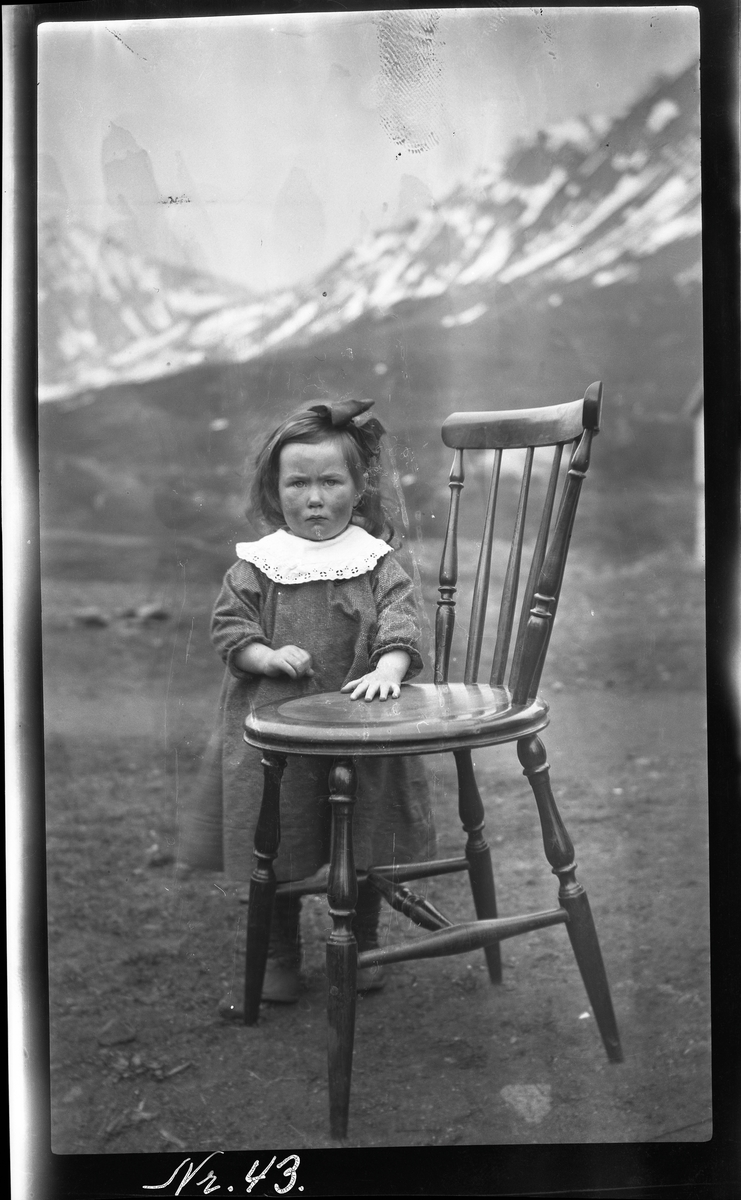 Portrett av liten pike stående ved stol utendørs.