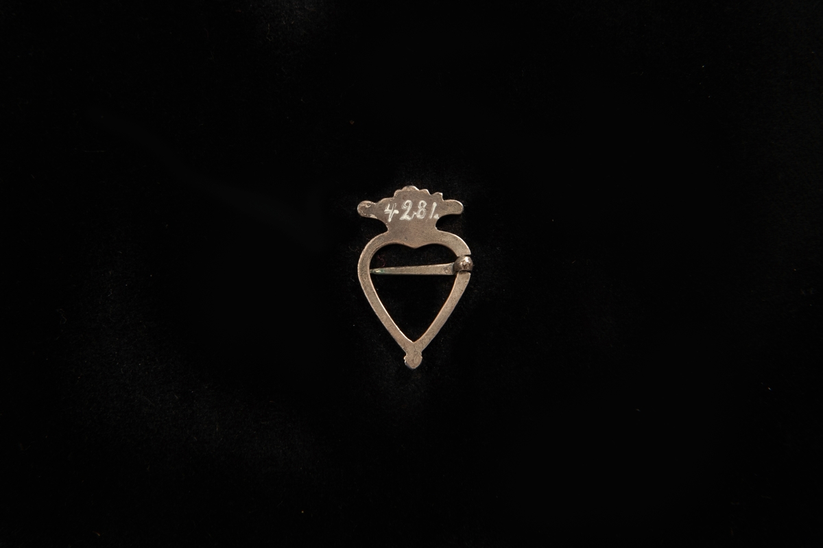 Hjärtformad sölja med krona överst som har graverat mönster i form av många små streck som går uppåt i kronans form. Söljan stängs med en nål. Består av metall som är silverpläterad(?).
