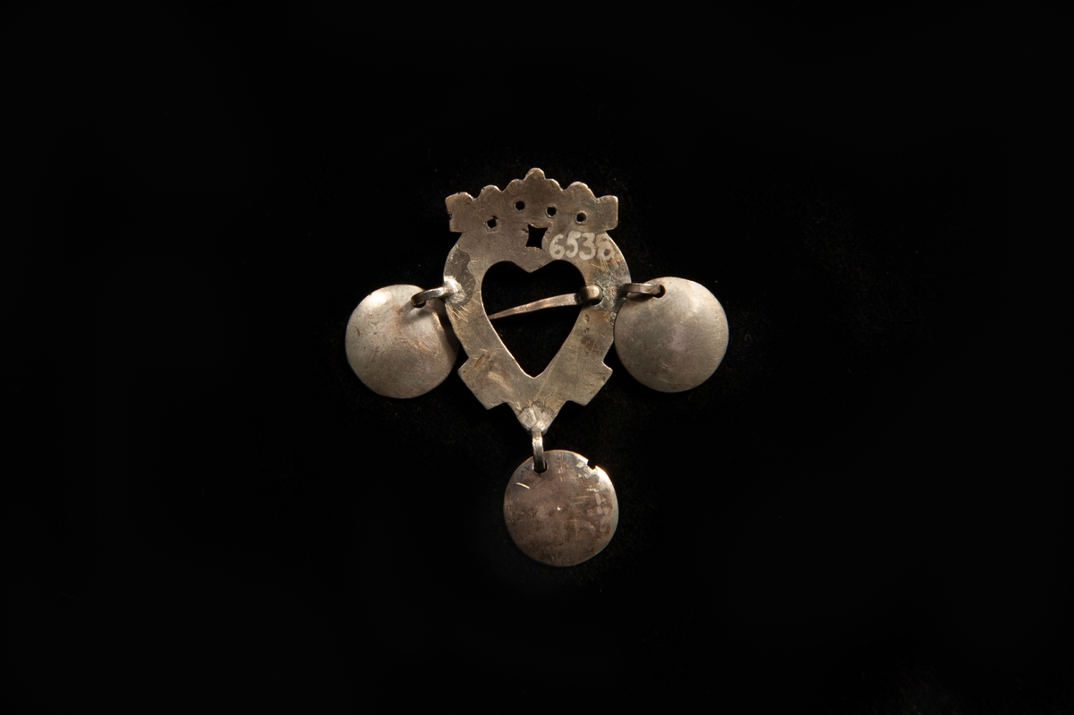 Hjärtformat spänne av graverat silver, med stiliserad krona. Tre släta skålar av silver hänger ner från spännet. Saknar stämplar.