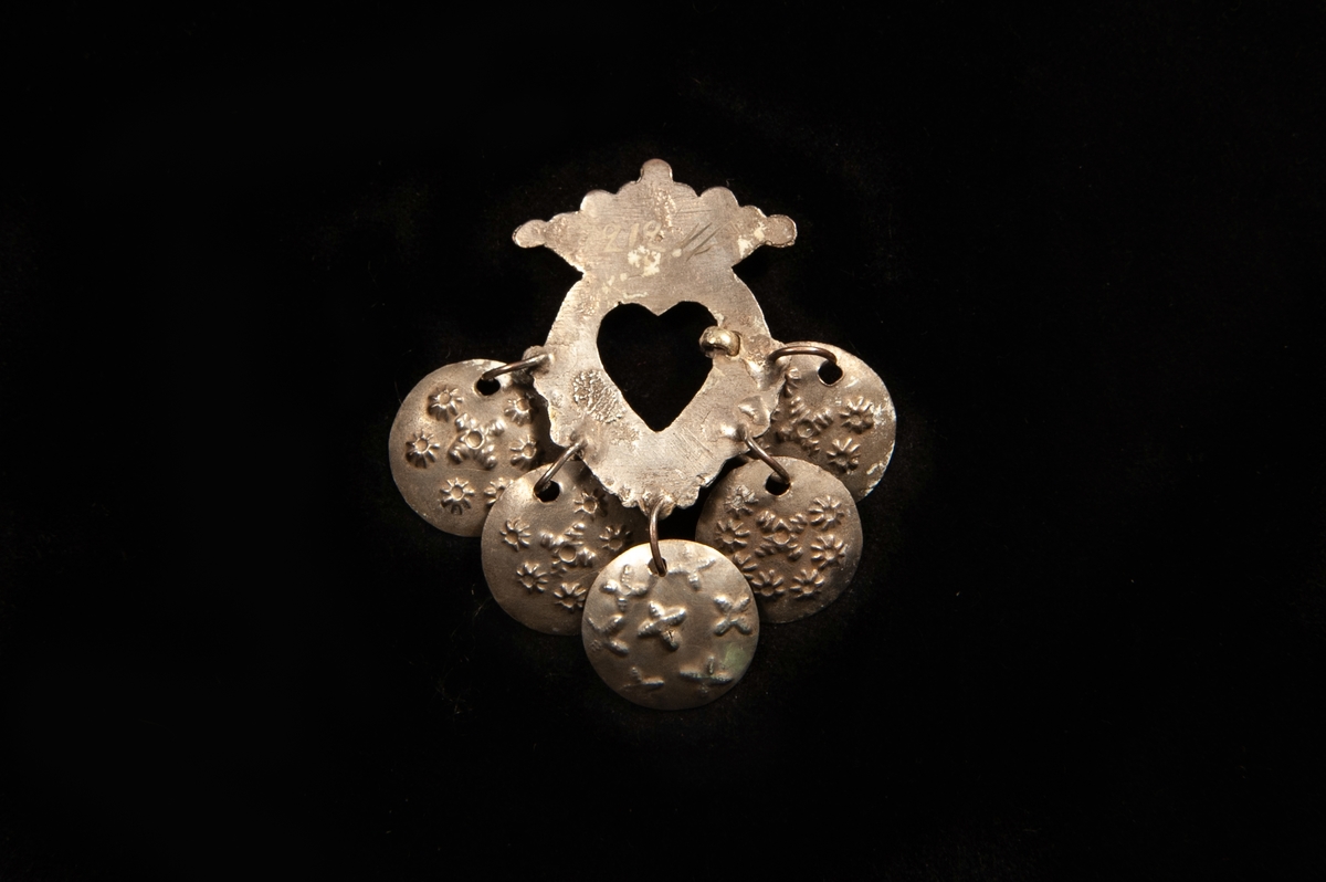 Hjärtformad bröstsölja av graverat och förgyllt silver, med nål. Krona överst och fem mönsterstämplade löv av silver hänger ner från spännet. Saknar stämplar.