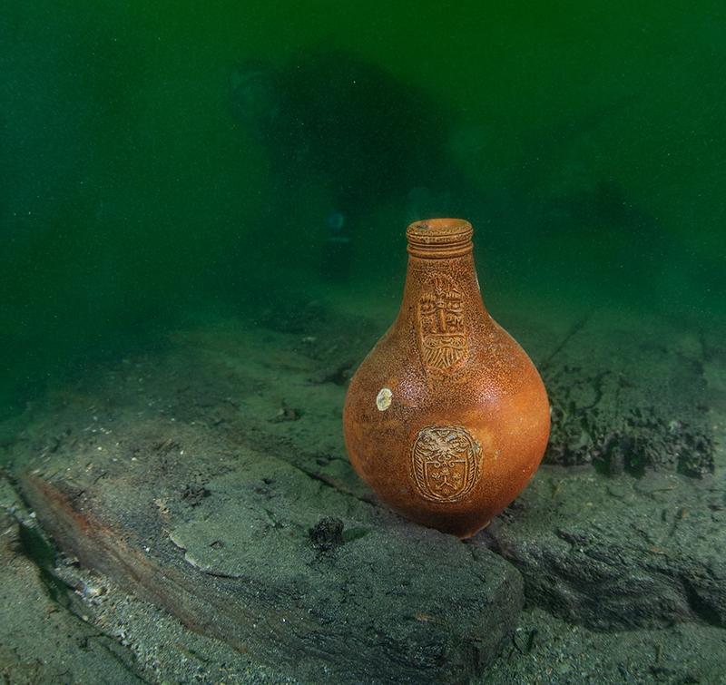 Bartmannskrukke i oransje keramikk, fra siste halvdel av 1600-tallet, står på bunnen blant båtdeler fra vraket av Pelikanen.