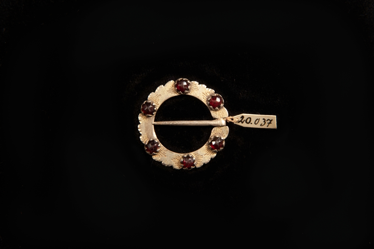 En rund ringsölja av silver med enkel graverad stråldekor runt sex små infattade stenar av rött glas. Uddig kant. Stämplad på baksidan.