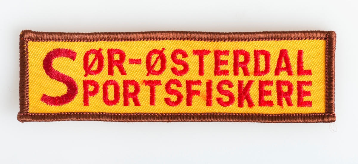 Rektangulært tøymerke som er brukt til egenprofilering av foreningen Sør-Østerdal Sportsfiskere. 
Teksten "Sør-Østerdal Sportsfiskere" er påsydd med rød tråd på et ornasjefarget stoff med brun kant som innramming. 