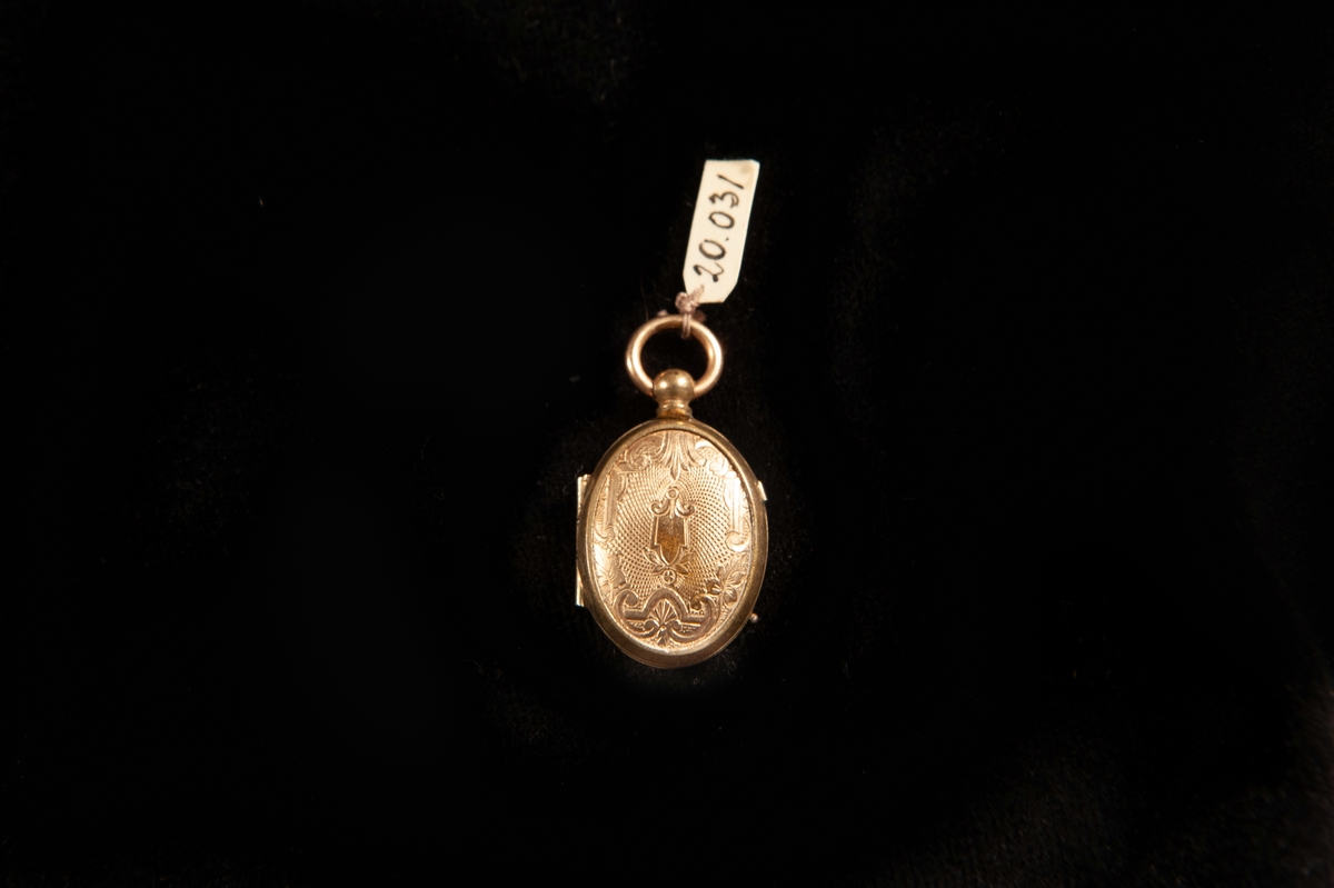 En oval medaljong i förgylld metall. Graverat mönster fram- och baktill i form av slingor m.m. i blandstil (nyrokoko/nyrenässans). Avsedd för två foton. Innanför ett av glasen finns en blond hårlock. Ostämplad.