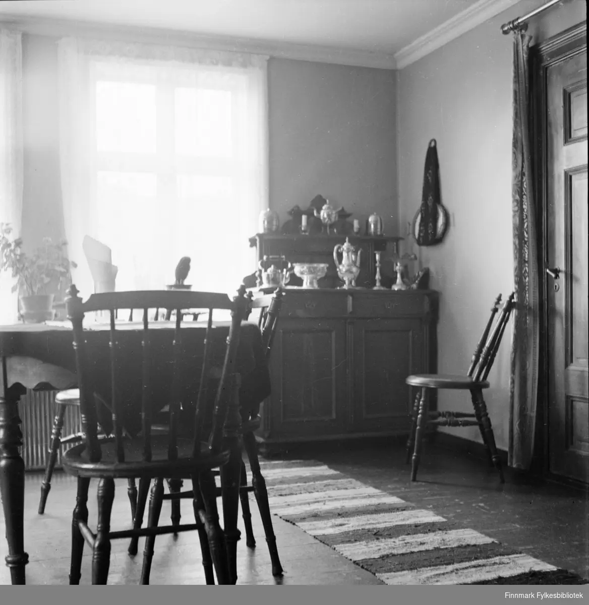 Interiør fra et hus i Kjøllefjord, fotografert i 1940. I forgrunnen sees et spisebord med dreide pinnestoler. I bakgrunnen sees et utskåret skatoll med sølvtøy servise oppstilt i flere nivåer. En fillerye ligger på gulvet.