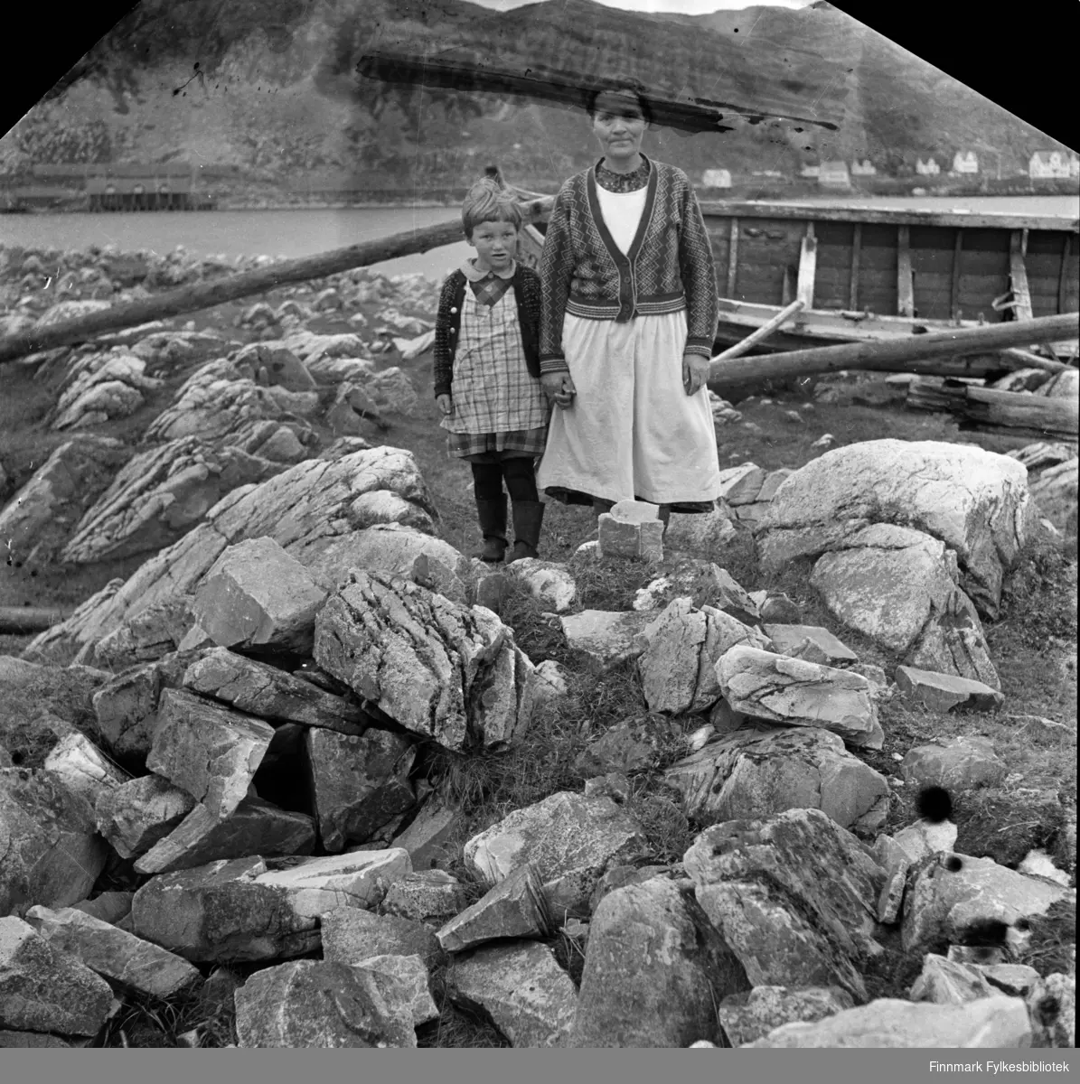 Ukjent kvinne med barn fotografert i Kjøllefjord, 1940. De står i et steinete landskap. En robåt ligger hvelvet på siden i bakgrunnen. Kvinnen har hvitt forkle under jakka, over kjolen. Det har også jenta som har rutete forkle over en rutete kjole. På beina har hun gummistøvler. I bakgrunnen ser man deler av Kjøllefjords bebyggelse over fjorden. Til høyre i bildet sees huset til kjøpmann og hurtigruteekspeditør Horst og til venstre for Horst sees Jon Svendsens hus.