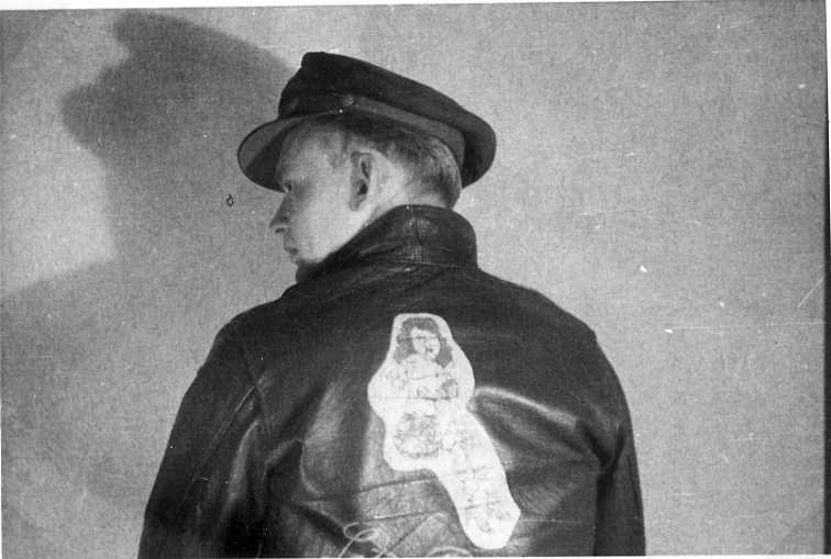 Troligen R G Fox, en ung man i uniform: skinnjacka med pinup-bild på ryggen, scarf och skärmmössa. Fotograferade bakifrån.