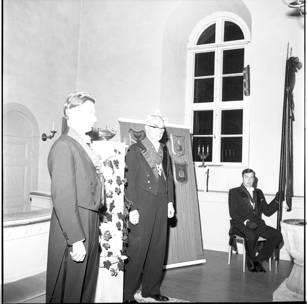 Sällskapet Par Bricole har sammankomst. Två män i högtidsdräkt står, sannolikt i Järstorps kyrkas absid.