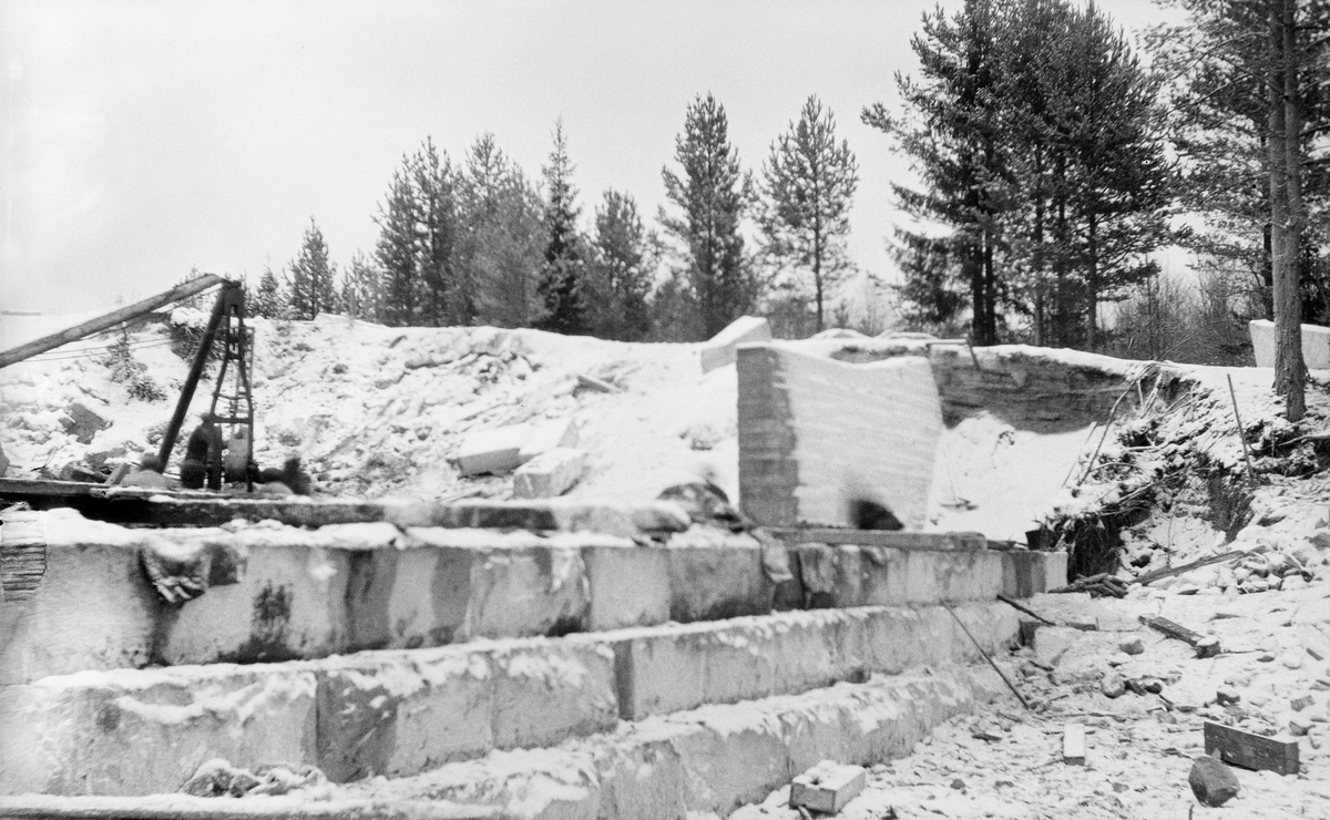 Kjellåsdammen i Flisavassdraget i Åsnes, fotografert mens dammen ble ombygd fra en steinfylt tømmerkistedam til en kvadersteinsmurt tørrmurkonstruksjon i perioden 1916-1924.  Fotografiet viser et trappetrinnsformet fundament av naturstein, som antakelig ikke var ferdigmurt da bildet ble tatt.  Muren føres mot en grusrygg – antakelig elvebakken – der det var gravd ut en del masse.  Så vel murverket som marka bakenfor var dekt av et tynt snølag da dette fotografiet ble tatt.  På elvebrinken i bakgrunnen vokste det furutrær.

Kjellåsdammen var en såkalt attholdsdam som kunne demme opp den nedre delen av Flisavassdraget i en lengde på opptil fem kilometer. Den dammen det avbildete anlegget avløste var bemannet med to damvoktere så lenge det pågikk fløting i Flisa. Tidlig på 1900-tallet var fløtingsledelsen i tvil om det svarte seg å holde seg med damanlegg og mannskap på dette stedet. Det ble snakket om at dammen samlet lite vann, og at det av den grunn kunne være aktuelt å rive hele Kjellåsdammen så snart den nedenforliggende delen av vassdraget var bedre «opprenset». I stedet ble det altså til at man etter rivinga av den gamle dammen bestemte seg for å bygge en ny. Den nye damkonstruksjonen ble reist i perioden 1916-24.

Den første dammen ved Kjellåsfossen ble oppført på midten av 1800-tallet - jfr. fanen «Opplysninger».