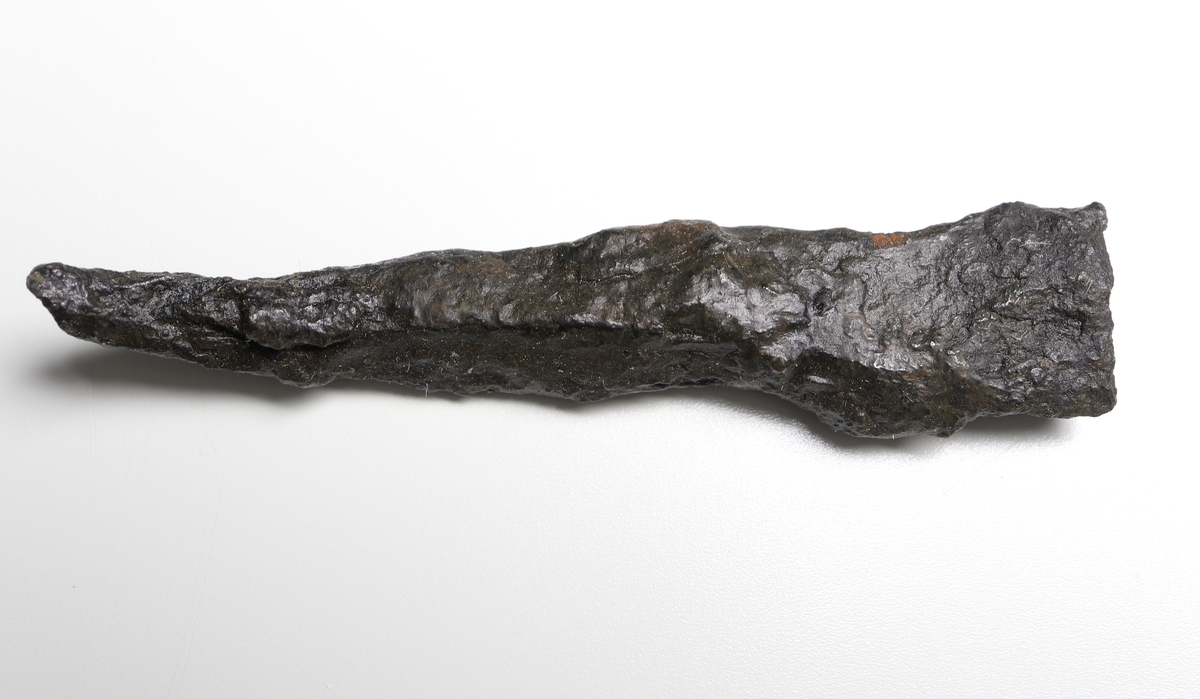 Fragment av en dolk eller annan spets med rombiskt tvärsnitt. Endast yttersta spetsen är bevarad. Vittrad och förböjd.