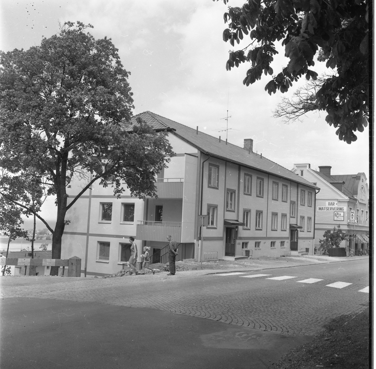 Nybyggt hyreshus på Brahegatan 1 i Gränna. Längre ner på gatan skyltas för Ritz Matsalar, mat och polkagrisar. En liten flicka skyndar efter sin mamma. En man i rutig skjorta tittar efter dem.