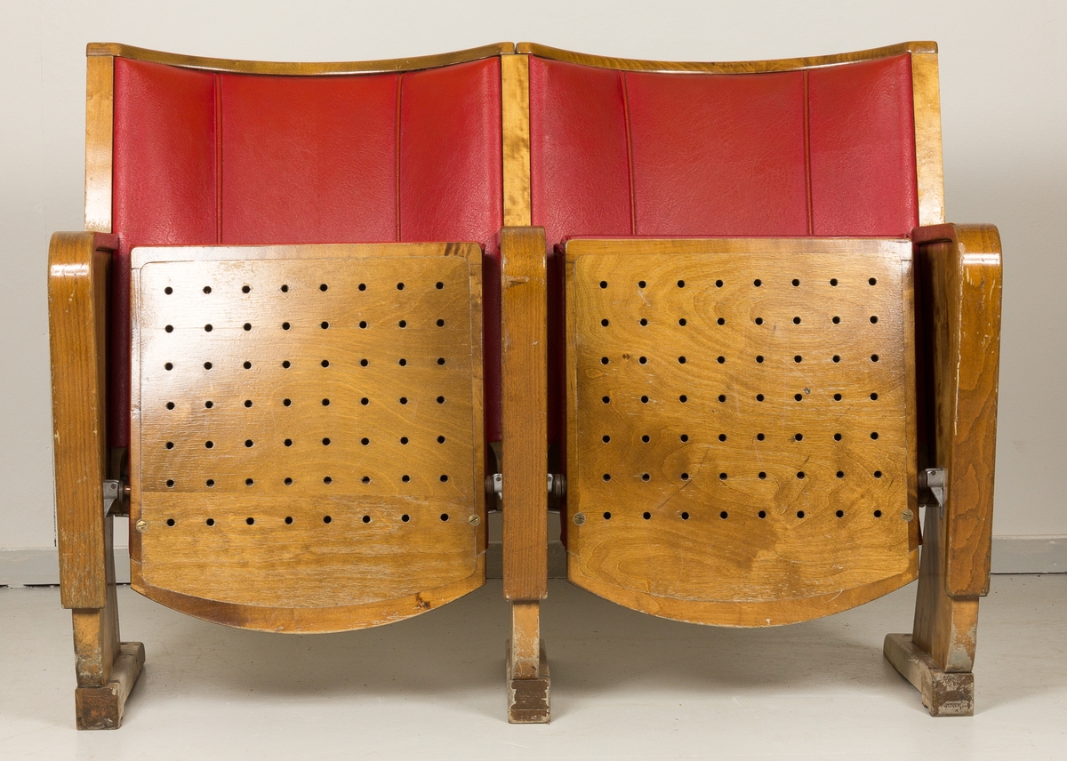 Kinostoler, to sammenhengende klappstoler i bjørk med armlener i alm, trukket i rødt skinn/skai. Brukt i Harstad kino fra 1954 til oppussingen i 1988.
