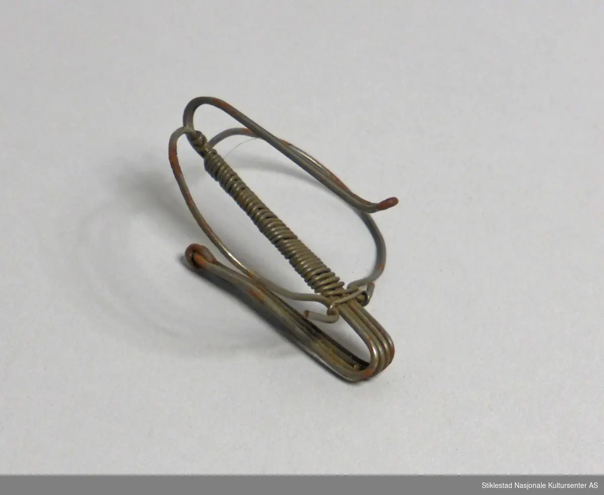 Nøstekrok laget av myk metalltråd. Formet to kroker fra enkel ring. Ene krok for å feste i jakke, forklekant eller lignende, den andre kroken for å hekte nøstet på. Hjemmelaget.