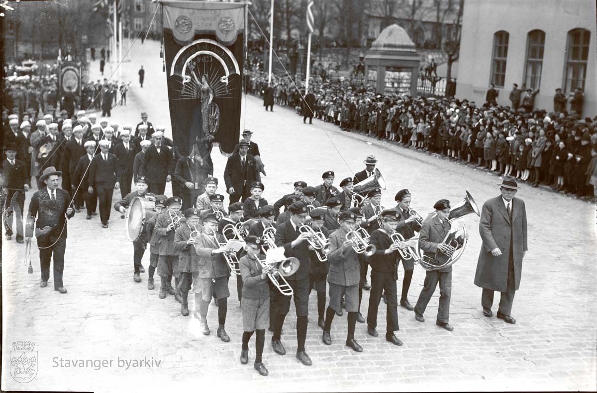 Et korps av unge gutter leder an foran fanen for Stavanger jern- & metallarbeiderforening. Tilskuere ved posthuset.