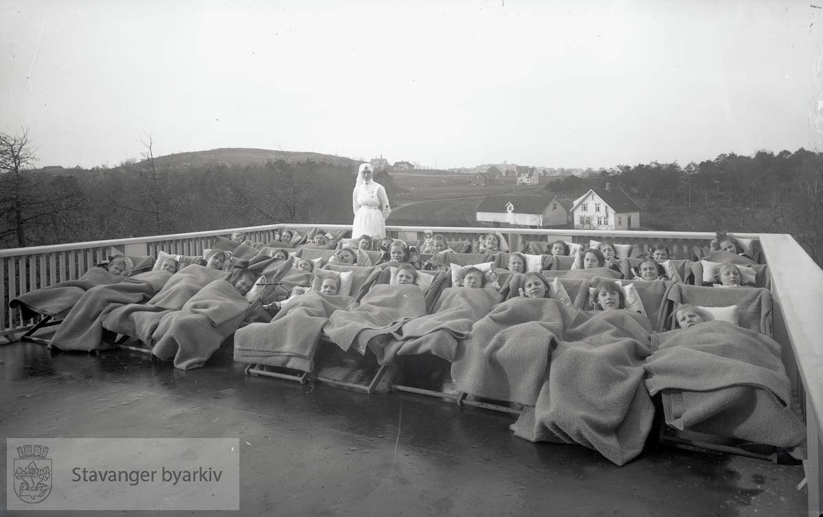 Friluftskolen for tuberkulosetruede barn var opprettet av Sanitetsforeningen i 1920. Den lå bare 200 m fra tuberkulosehjemmet i Ramsvik / Ramsvig. Jenter i liggestoler, godt dekker av ulltepper, på luftealtanen til tuberkulosehjemmet. I bakgrunnen til venstre er Varden. Fjernt i bakgrunnen i vest skimtes Våland.