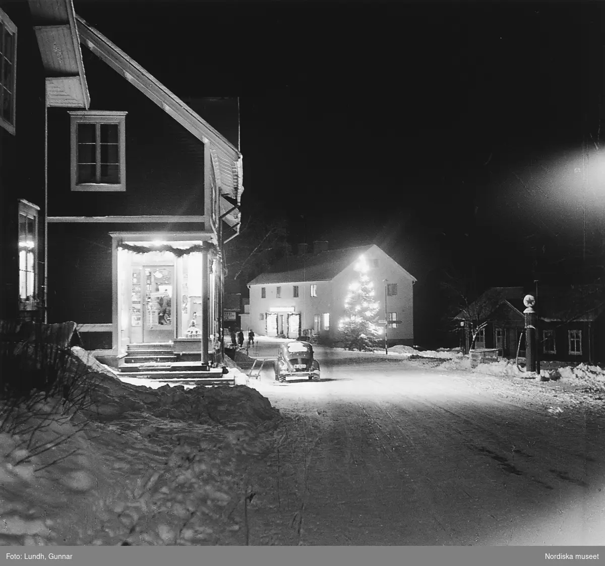 Nattbild på en snötäckt gata i Tällberg år 1950. En bil står parkerad utanför en affär. På andra sidan gatan en bensinpump. En stor julgran med belysning står vid gatan.