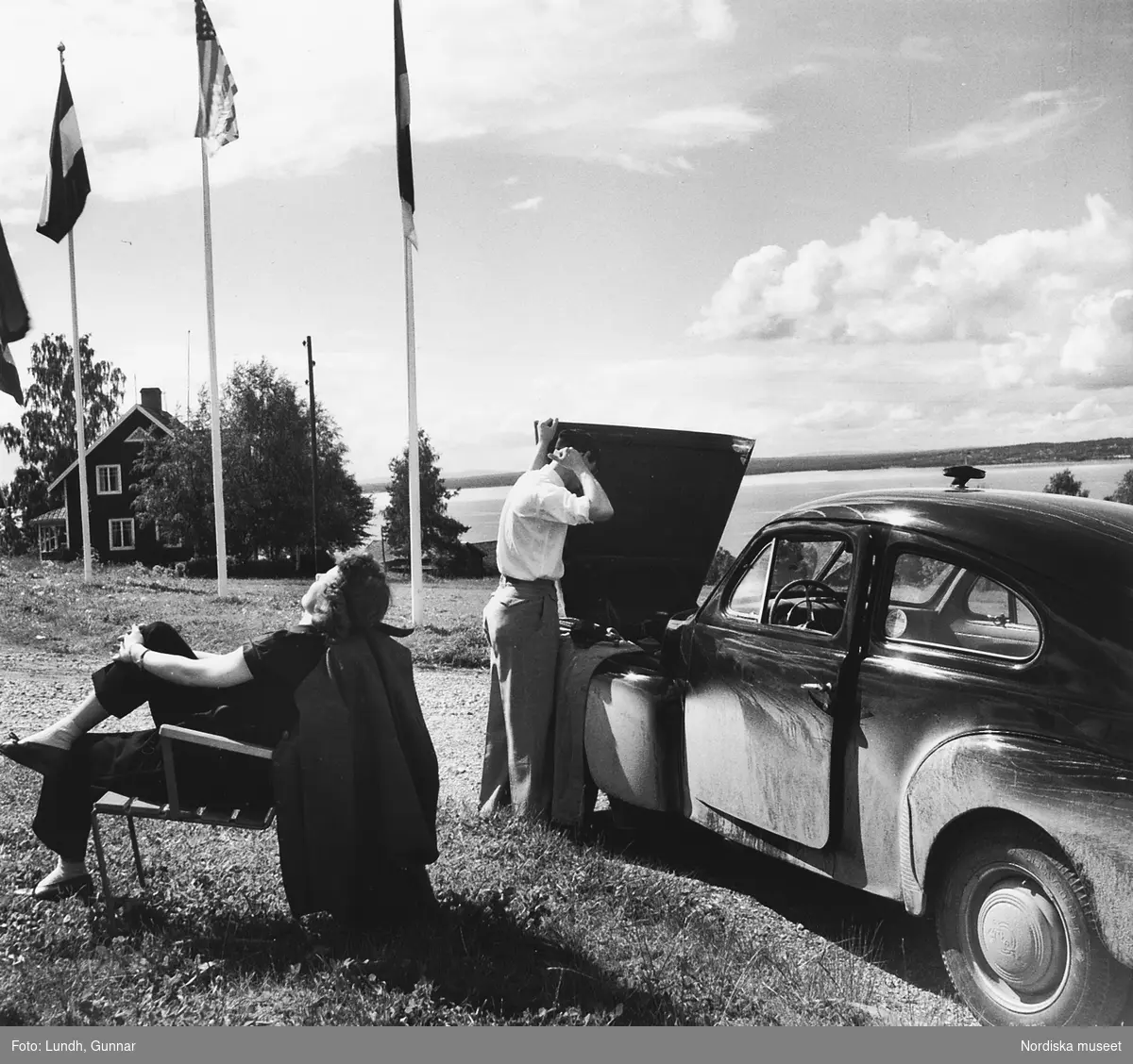 En kvinna sitter i en stol och solar medans en man står vid en öppen motorhuv på en Volvo PV årsmodell 1951. I bakgrunden flaggstänger och ett hus. Bilden tagen i Rättvik år 1952.