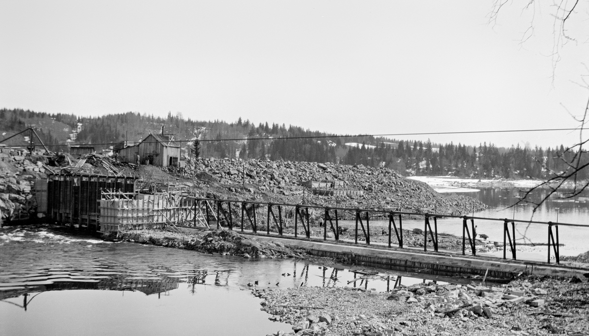 Bygging av nåledam - såkalt «fermettedam» - ved Vormas østre bredd ved Svanfossen i Fenstad i Nes kommune på Øvre Romerike, antakelig fotografert tidlig på våren, før vannstanden i elva begynte å stige.  Den øvre delen av dette vassdraget ble noe regulert i slutten av 1850-åra, for å gjøre det mulig å kople den da nyetablerte hovedbanen med dampskipsruter på Mjøsa.  Den første dammen ved Svanfossen skal ha vært bygd i 1910.  Her ser vi hvordan elva faller over en terskel på elvebotnen.  På den østre delen av denne ble det montert en såkalt «fermettedam», en stålkonstruksjon som skulle bære ei gangbru, der fløterne kunne gå når de skulle «sette dammen» (stenge) eller «sprette» (åpne) den. Dette ble gjort ved hjelp av «nåler», plank som ble stukket mot strømmen, slik at den lå an mot en terskel i botnen av elveløpet og mot gangbrua.  Disse nålene ble satt tett i tett, slik at de sammen dannet en vegg mot vannet.  Når dammen ble åpnet, brukte karene små kjetting- eller taustubber med ei jernnål i enden, som ble tredd gjennom hull i den øvre delen av nålene, og trakk til. Til venstre i bildet ser vi hvordan anleggsarbeiderne hadde forskalet kar til et lukeløp, som seinere skulle få en overbygning, et hus, som lukene kunne reguleres fra (jfr. bl. a. SJF.1989-02451 og SJF.198902452). Informasjon i Glomma fellesfløtingsforenings arkiv indikerer at dette fotografiet skal være tatt i 1916. Vi er ikke sikre på at dette er riktig, for bygginga av denne dammen skal ha foregått 8-9 år tidligere - jfr. fanen «Opplysninger».
