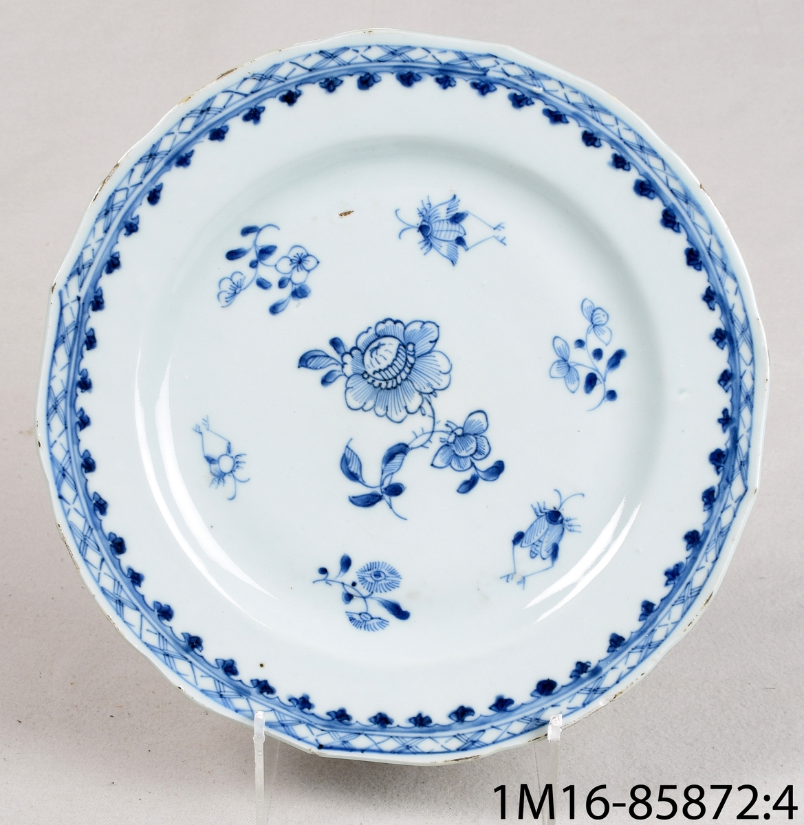 Tallrik av vitt porslin med svagt vågig kant. Har handmålad blå dekor föreställande blommor och insekter. Har dekor både på ovansida och undersida.