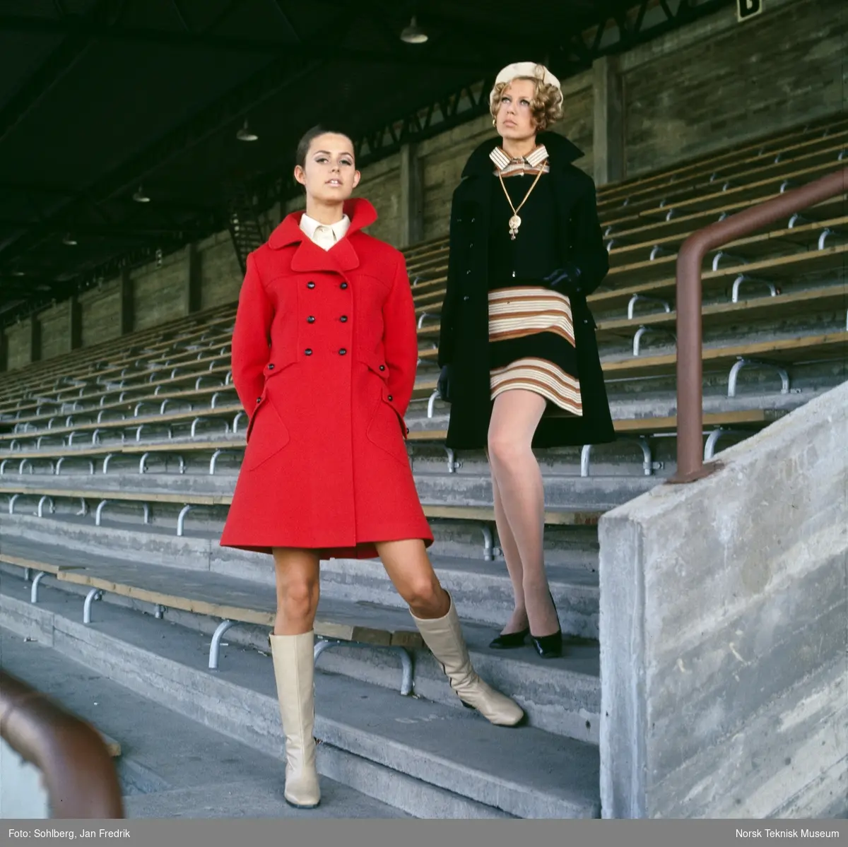 Motebilde, to kvinner, den ene i mørk kåpe over stripete kjole, den andre i rød kåpe, på tribune på idrettsplass. Bildet er en del av en serie der noen er publisert i ukebladet Det Nye 1969, nr. 38.