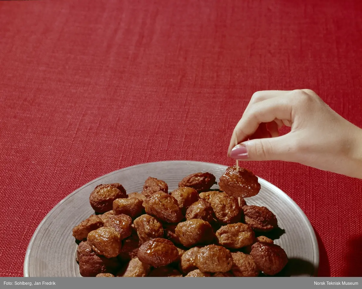 Reklamebilde for kjøttboller av typen Bolls. Hermetisk norsk bondekost. Kjøttbollene ligger på en tallerken, en kvinnehånd forsyner seg med en kjøttbolle ved hjelp av en tannpirker.