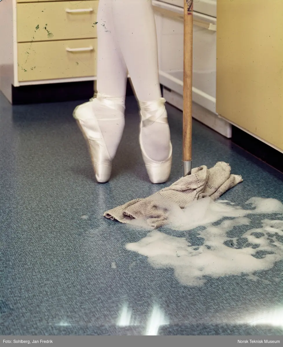 Mote- eller reklamefotografi av føttene til en person i tåspissko. Vedkommende befinner seg på et kjøkken og er i ferd med å vaske gulvet.