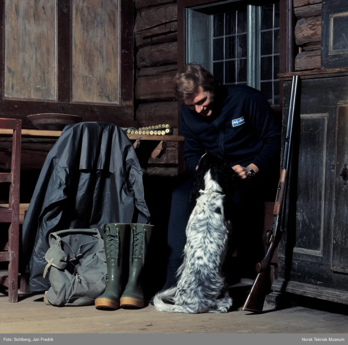 Tidlig norsk motefotografi. Mannlig modell viser fram jaktantrekk. Ved siden av ham står jaktgeværet klart. Han klapper en hund, engelsk setter.