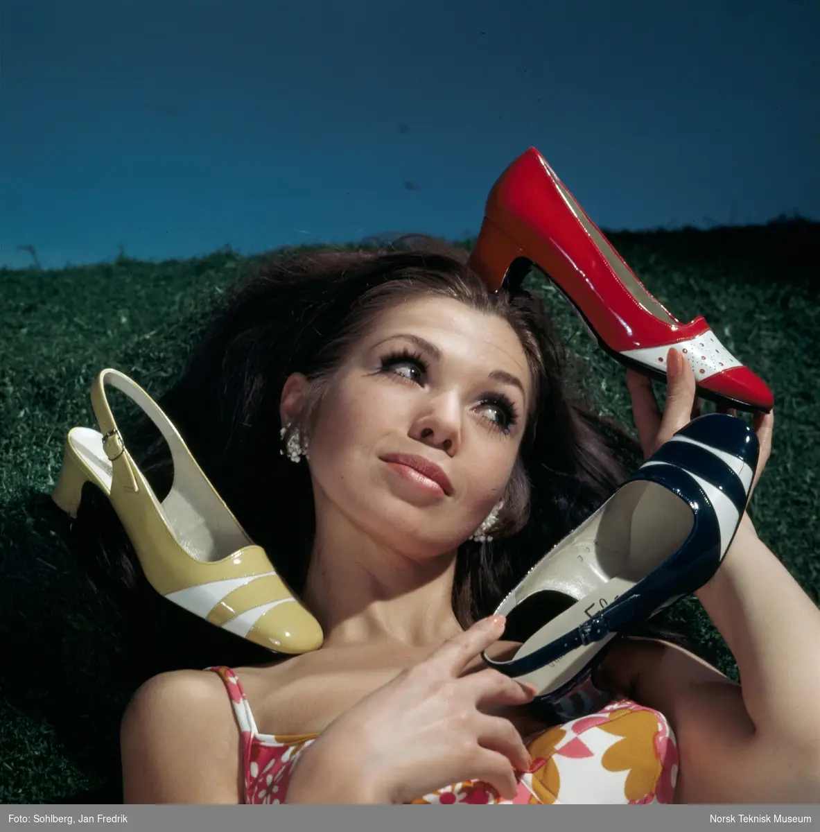 Tidlig norsk reklame- / motefoto. En kvinnelig modell ligger i gresset med flere sko rundt seg. Hun virker fornøyd med tidens skomote.