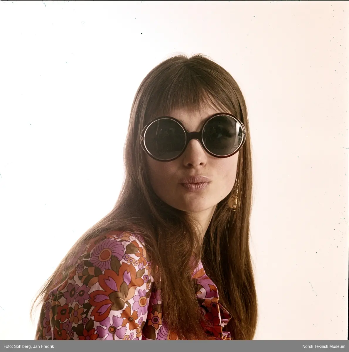 En kvinnelig modell viser fram tidens solbrillemote. Hun har langt hår hengende løst. Hun har store, runde solbriller og bluse med mye mønster med rosa og oransje blomster. Hippie-stil.