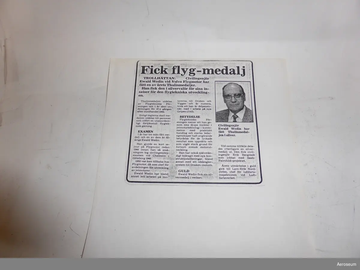En grå och svart pärm med ett färgfoto i träram. Fotot föreställer Evald Wedin framför en jetmotor, troligen RM6. Med fotot föjer ett kopierat tidningsurklipp och ett årsmötesprotokoll från Flygtekniska Föreningen från 1985 där det meddelas att Evald Wedin tilldelats Thulinmedaljen.