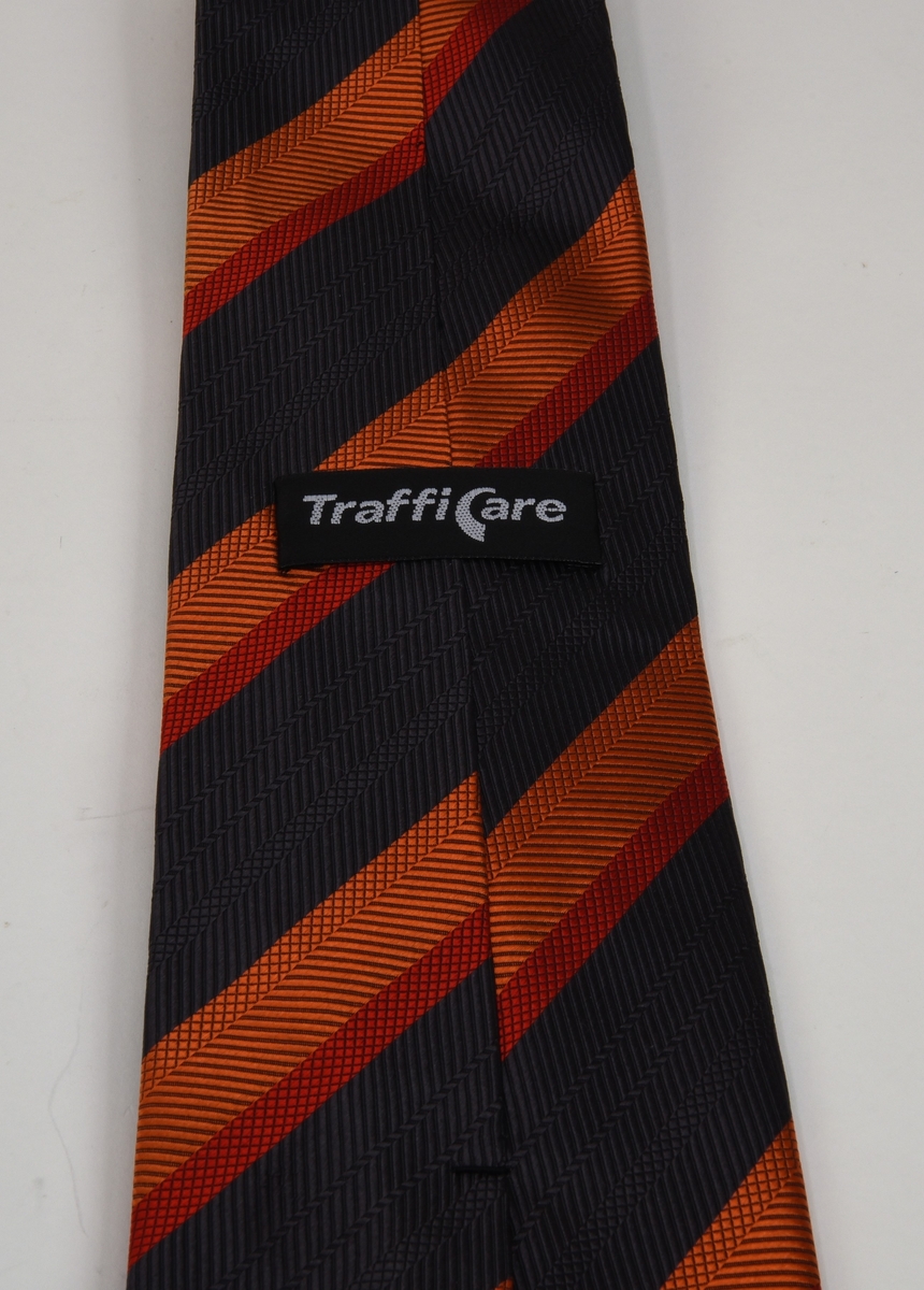 Slips med breda diagonala ränder i svart, röd och orange. Slipsen är tillverkad av siden och är rand-och rutmönstrad samt fodrad med en mörkblå textil.